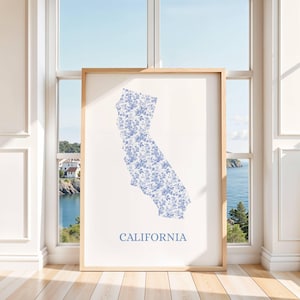 May include: A framed print featuring the outline of California filled with a blue floral pattern. The word "CALIFORNIA" is printed in a matching blue font below the state outline. The print is displayed in a room with a window overlooking a coastal view.