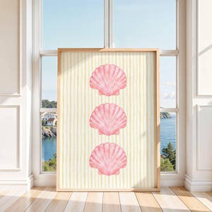 May include: A rectangular wall art print in a light wooden frame featuring three vertically aligned pink scallop shells centred on a background of thin, pale yellow vertical stripes. The artwork is displayed against a bright window backdrop.