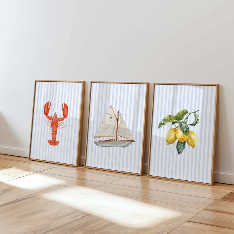 Op de afbeelding: Drie ingelijste aquarelprints met nautische thema's. E&eacute;n toont een rode kreeft, een andere een zeilboot en de derde citroenen op een tak. Elke print heeft een blauw-wit gestreepte achtergrond en een houten lijst.