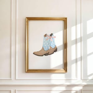 May include: A framed print of a cowboy boot. The boot is tan with a light blue shaft adorned with white stars. A red, white, and blue bow decorates the top. The frame is gold, set against a white wall.