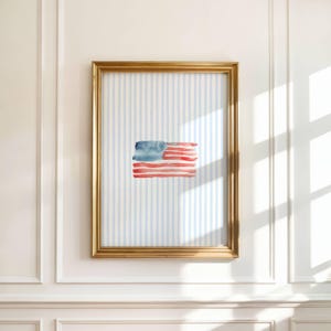 May include: A framed watercolor painting of the American flag. The flag is depicted with red and blue brushstrokes, set against a light blue and white striped background. The frame is gold.
