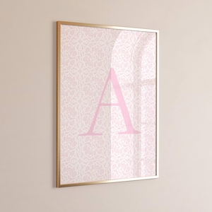 May include: A framed print displaying the letter "A" in pink, set against a patterned backdrop. The artwork is enclosed within a gold-toned frame. The background features a delicate, repeating floral pattern in a pale pink.