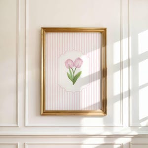 Op de afbeelding: Een ingelijste bloemenprint met twee roze tulpen met groene bladeren. Het kunstwerk is geplaatst tegen een roze en witte gestreepte achtergrond, omlijst in een goudkleurige lijst. De print is op een witte muur gemonteerd.