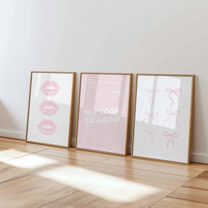 Peut inclure: Trois impressions d'art encadrées avec des cadres dorés. L'impression de gauche présente trois contours de lèvres roses. L'impression centrale contient le texte "Good Check" et "YOU LOOK SO GOOD!" sur fond rose. L'impression de droite affiche des illustrations de nœuds roses.