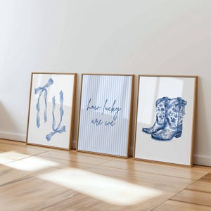 Puede incluir: Tres impresiones artísticas enmarcadas con diseños de acuarela. Las impresiones presentan una cinta azul, el texto "how lucky are we" y un par de botas de vaquero azules con motivos florales. Los marcos tienen un acabado de madera clara.