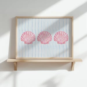 May include: A framed print featuring three pink scallop shells on a light blue and white striped background. The artwork is displayed on a wooden shelf against a white wall, with natural light casting shadows.