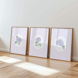 May include: Three framed art prints featuring watercolor illustrations of purple hydrangea flowers. Each print has a light purple and white striped background and a wooden frame. The hydrangea is set in a white, scalloped-edge oval.