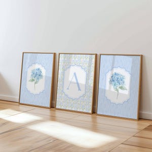 May include: Three framed wall art prints with light wood frames. The outer prints feature blue hydrangea illustrations on a light blue and white striped background. The center print displays a large blue letter A on a floral patterned background.