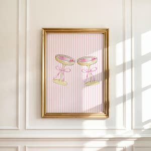 May include: A framed print showing two champagne glasses with pink liquid, decorated with pink bows. The background has a pink and white striped pattern, within a gold frame. The artwork is displayed on a white wall.