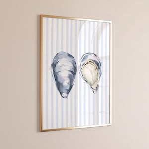 May include: Watercolor print of two oysters in a gold frame. One oyster is closed, the other is open, revealing a creamy interior. The background features vertical blue and white stripes, creating a coastal theme. The artwork is framed with a gold border.