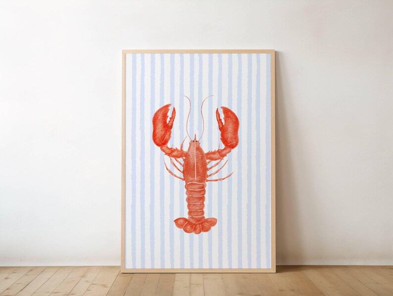 Op de afbeelding: Een ingelijste kunstdruk met een aquarel illustratie van een rode kreeft. De kreeft staat tegen een achtergrond van verticale blauwe en witte strepen. Het kunstwerk wordt gepresenteerd in een licht houten frame.
