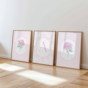 Peut inclure: Trois cadres muraux avec des bordures en bois clair. Les deux cadres extérieurs présentent des illustrations d'hortensias roses sur un fond rayé blanc et rose. Le cadre central affiche une grande lettre A rose dans un encadrement décoratif.