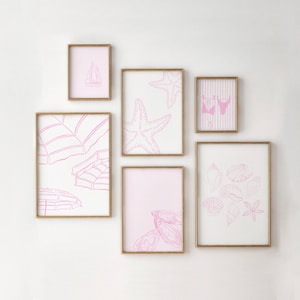 May include: A collection of six framed art prints with gold frames. The prints showcase pink line drawings of beach themes, such as a sailboat, starfish, beach umbrellas, seashells, and a bikini. The art is mounted on a white wall.
