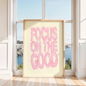 May include: A framed art print with the words "FOCUS ON THE GOOD" in pink bubble letters. The artwork has a light yellow background and a wooden frame. The print is displayed in front of a window overlooking a coastal scene.