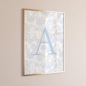 Pode incluir: Arte de parede emoldurada com um padrão floral azul claro e uma grande letra "A" azul claro. A obra de arte está emoldurada em uma moldura dourada, criando uma peça decorativa.