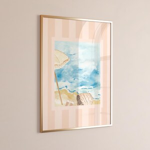 Puede incluir: Impresi&oacute;n en acuarela enmarcada de una escena de playa con una sombrilla beige, oc&eacute;ano azul y arena. La obra de arte est&aacute; en un marco dorado y sobre un fondo a rayas rosas y blancas. El tema es la decoraci&oacute;n costera.