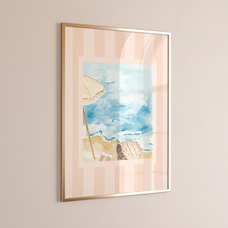 Puede incluir: Impresi&oacute;n en acuarela enmarcada de una escena de playa con una sombrilla beige, oc&eacute;ano azul y arena. La obra de arte est&aacute; en un marco dorado y sobre un fondo a rayas rosas y blancas. El tema es la decoraci&oacute;n costera.