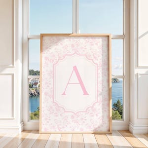 May include: A rectangular wall art print in a light wooden frame featuring a pale pink floral pattern background. Centered on the print is a white scalloped-edge frame containing a large, bold, capital letter A in a soft pink color.