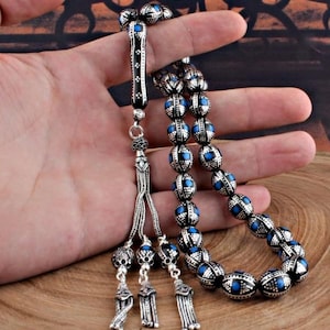 Könnte beinhalten: Gebetskette in Silber mit blauen Akzenten. Die ovalen Perlen sind aufgefädelt und weisen detaillierte Muster auf. Quaste und Verbindungsstücke sind ebenfalls silberfarben, wodurch ein einheitliches Design entsteht. Die Gebetskette wird in einer Hand gehalten.