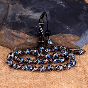Può includere: Una collana di perline di preghiera nera con dettagli argentati, punti rossi e blu e un manico decorativo. Le perline ovali sono infilate con un cordino nero. Lo sfondo è una superficie scura e strutturata.