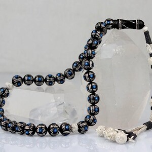 Può includere: Una collana di perline di preghiera nere e argentate con dettagli blu. Le perline rotonde sono decorate con motivi, con un nappa argentata e terminali decorativi. La collana è esposta su uno sfondo cristallino trasparente.