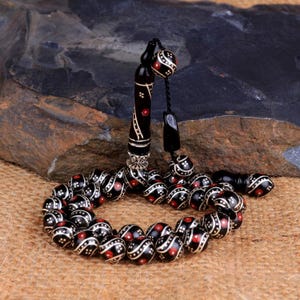 Peut inclure: Un chapelet noir et rouge. Les perles rondes sont ornées de détails blancs et rouges. Le pompon et la poignée sont noirs avec des détails blancs et rouges. Les perles sont présentées sur une surface brune.
