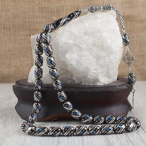 Può includere: Una collana di perline di preghiera nera e argento con dettagli blu. Le perline hanno un design a spirale e sono collegate da una catena d'argento. La collana è esposta su un supporto di legno con una roccia bianca sullo sfondo.