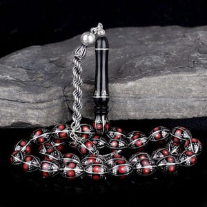 May include: A black and silver prayer bead set with red detailing. The oval beads feature a silver pattern and red central dots. The tassel and chain are silver. The prayer beads are presented on a dark grey stone surface.