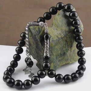 May include: A black beaded prayer bead necklace with a silver-colored chain and accents. The beads are round and glossy. The pendant has the word "Bingöl" in white lettering. The necklace is displayed against a green and brown stone.