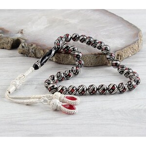 May include: A beaded prayer rosary with black and red beads, accented with white detailing. The rosary features a black and silver handle and a silver tassel. The background includes a white and grey stone.