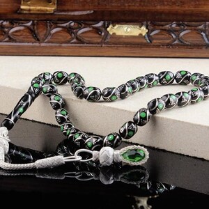 Può includere: Una collana di perline di preghiera nere e verdi con accenti argentati. Le perline hanno un motivo decorativo con dettagli bianchi e verdi. Una gemma verde si trova alla fine della collana. Lo sfondo include una scatola di legno.