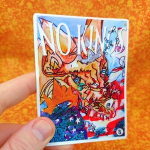 May include: A square sticker with the words "NO KINGS" in white block letters. The sticker features a colorful, abstract illustration with orange, blue, red, and white elements. The sticker is held in a hand against an orange background.
