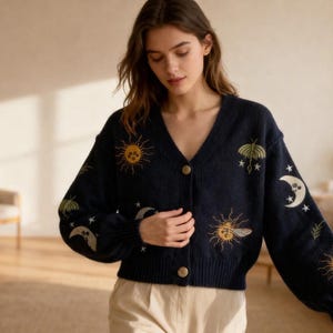 Peut inclure: Cardigan bleu marine avec broderies de motifs célestes : soleils, lunes et étoiles. Le pull à col en V a une fermeture boutonnée et des manches longues. Le modèle porte un pantalon beige.