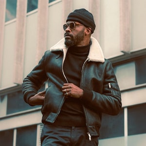 Op de afbeelding: Een zwarte beanie en zonnebril worden gedragen met een zwarte coltrui en een donkergrijze bomberjack met een witte schapenvachtkraag. De jas heeft een ritssluiting en een zak op de linkermouw.