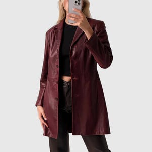 May include: A long, burgundy leather coat with a button-up front and a classic lapel collar. The coat appears to be knee-length and is worn over a black top and black pants. The coat has long sleeves and a tailored fit.