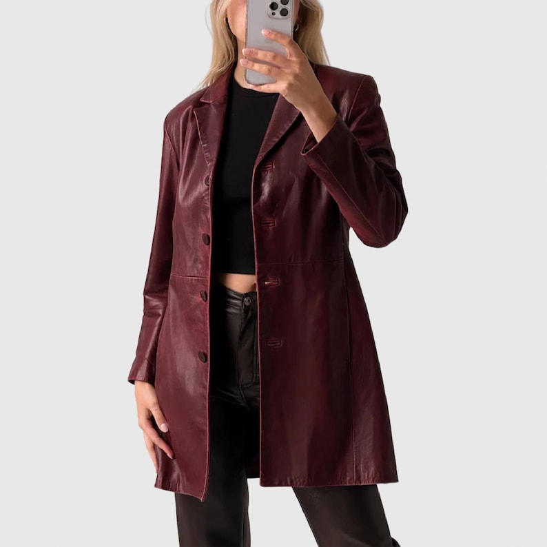 May include: A long, burgundy leather coat with a button-up front and a classic lapel collar. The coat appears to be knee-length and is worn over a black top and black pants. The coat has long sleeves and a tailored fit.
