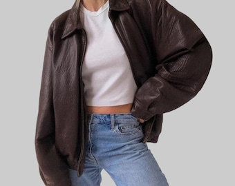 May include: A brown leather jacket with a collared neckline and a zipper closure. The jacket is worn over a white t-shirt and blue jeans. The jacket has a relaxed fit and a slightly oversized look.