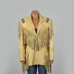 May include: A tan suede jacket with long fringe detailing across the chest, shoulders, and sleeves. The jacket features decorative stitching and embellishments on the shoulders and sleeves. It has a classic collar, two front pockets, and a button-up closure.