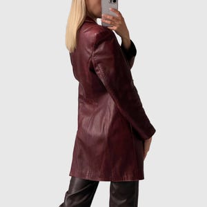 May include: A burgundy leather blazer with a classic lapel and long sleeves. The blazer has a tailored fit and falls to mid-thigh. The garment is paired with dark brown leather pants. The overall style is vintage and chic.