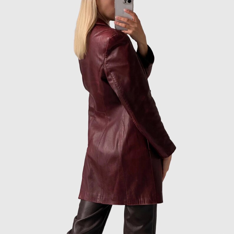 May include: A burgundy leather blazer with a classic lapel and long sleeves. The blazer has a tailored fit and falls to mid-thigh. The garment is paired with dark brown leather pants. The overall style is vintage and chic.