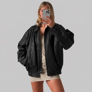 May include: An oversized black leather jacket featuring a classic collar, long sleeves with buttoned cuffs, and a front zipper closure. The jacket has a textured finish, a relaxed fit, and an elasticized hemline, worn over a simple beige mini dress.