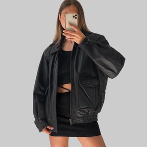 Può includere: Giacca bomber in pelle nera con chiusura a zip, due tasche frontali e orlo a coste. La giacca ha un colletto classico e polsini con bottoni. La modella indossa un top e una gonna neri.