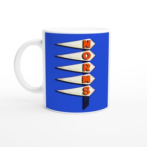 May include: White ceramic mug with a blue background featuring the word "NORMS" in orange letters. The letters are arranged vertically on white arrow-shaped signs. The mug has a white handle.