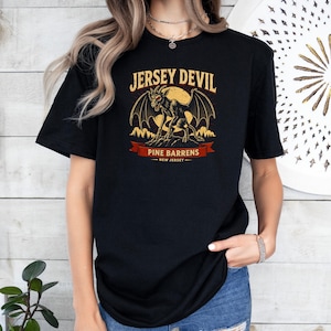 Può includere: Una t-shirt nera a maniche corte con una stampa grafica del Jersey Devil alato, in piedi su una roccia davanti a una luna gialla. Il testo recita JERSEY DEVIL, PINE BARRENS e NEW JERSEY su uno stendardo rosso e sotto.