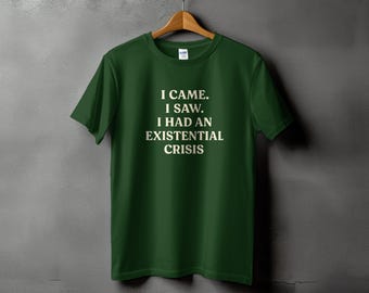 May include: A dark green t-shirt with the text "I CAME. I SAW. I HAD AN EXISTENTIAL CRISIS" in white, hanging on a wooden hanger against a gray background. The shirt is a crew neck style.