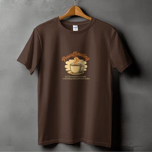 Puede incluir: Camiseta marrón con un diseño temático de café. El diseño presenta una taza de café con vapor y la palabra "Procaffinating" encima. Debajo de la taza está la frase "la tendencia a retrasar todo hasta que hayas tomado café."
