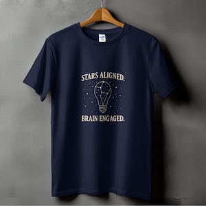 Può includere: T-shirt blu navy con la scritta bianca "STARS ALIGNED. BRAIN ENGAGED.". Il design presenta una forma di lampadina con un motivo a costellazione all'interno. La maglietta è appesa a una gruccia di legno.