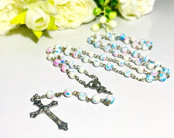 Könnte beinhalten: Ein Rosenkranz mit weißen, mit rosa und blauen Farbtupfern versehenen Perlen, auf einer silbernen Kette aufgereiht. Der Rosenkranz hat ein silbernes Kruzifix und ein dekoratives Mittelstück. Weiße Blumen sind im Hintergrund.