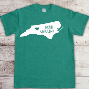 以下が含まれることがあります： ターコイズグリーンのTシャツに、ノースカロライナ州の白いアウトラインと「NORTH CAROLINA」の文字が書かれており、テキストの左側に小さなハートがあります。