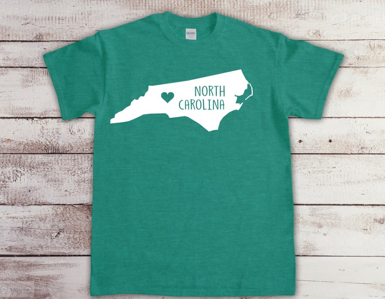 以下が含まれることがあります： ターコイズグリーンのTシャツに、ノースカロライナ州の白いアウトラインと「NORTH CAROLINA」の文字が書かれており、テキストの左側に小さなハートがあります。